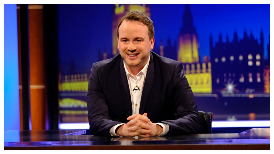 Man sitting at a desk in front of a blurred background of the Houses of Parliament.