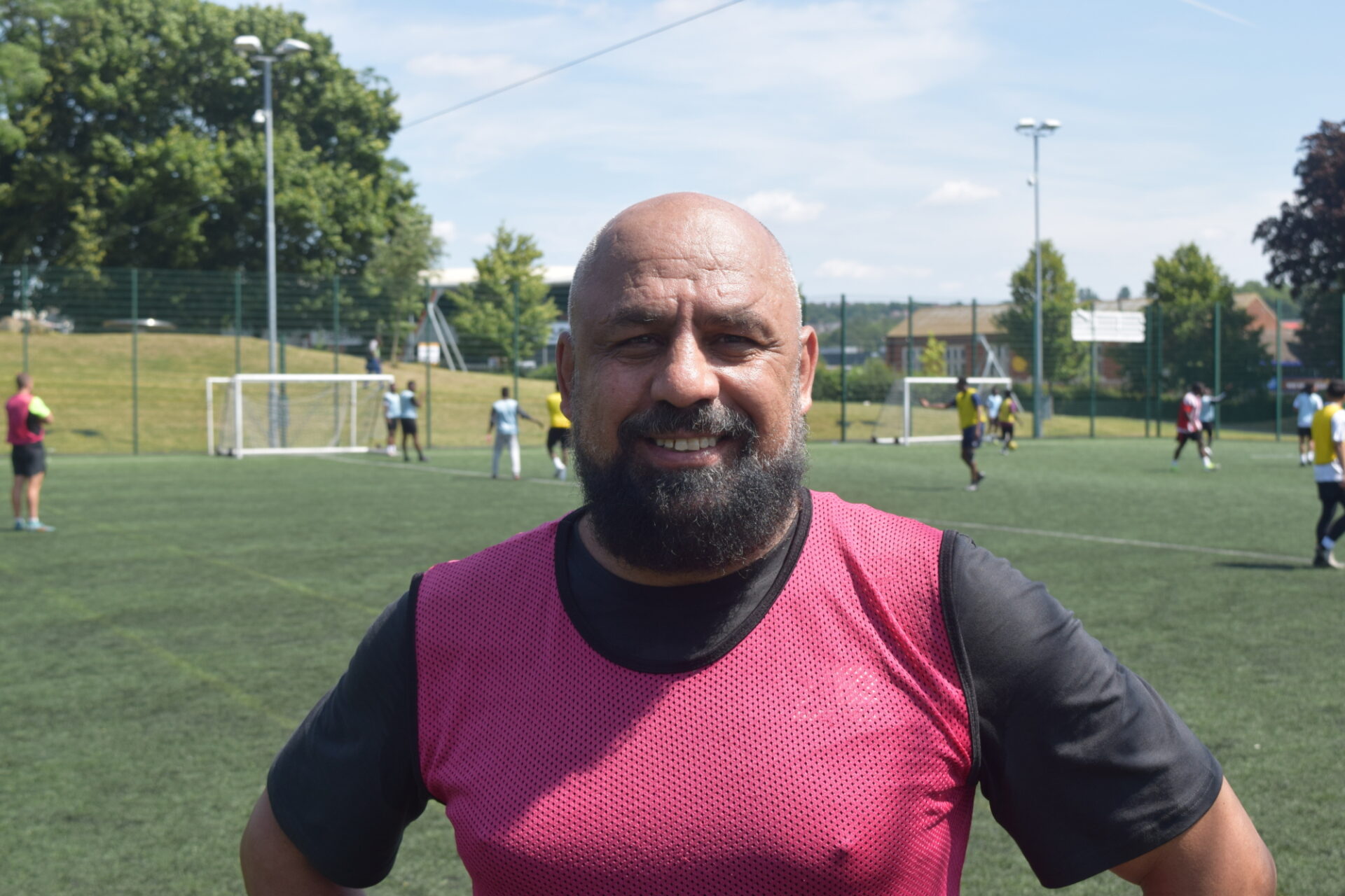 A middle-aged man with a beard stands confidently on the side of a football pitch, hands on his hips.