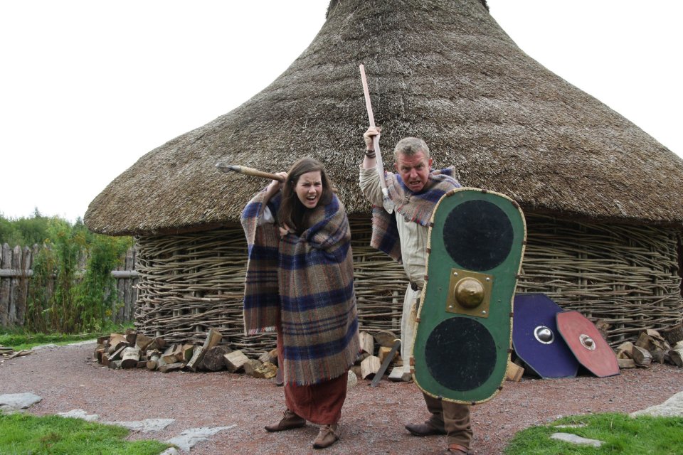 Two people in Celtic attire reenacting a scene at an Iron Age dwelling reconstruction.