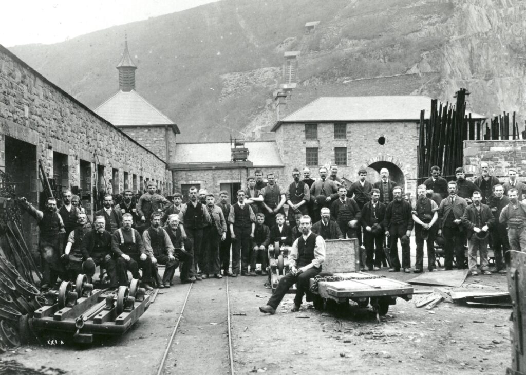A large group of men, likely quarry workers, pose together outside stone industrial buildings with carts and equipment on tracks, set against a backdrop of hills and a quarry.