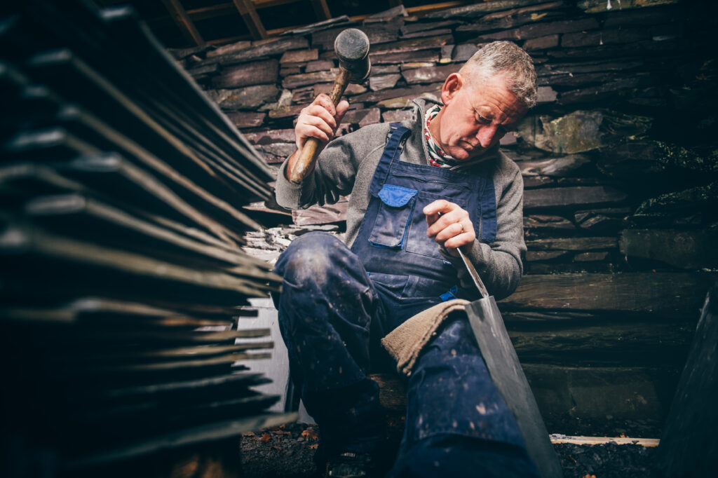 A man wearing blue overalls uses a hammer and chisel to shape a piece of stone inside a workshop, with stacked slabs of stone in the foreground and a stone wall behind him.