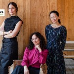 Three women, one seated, in front of a rustic yet refined wood panel wall.