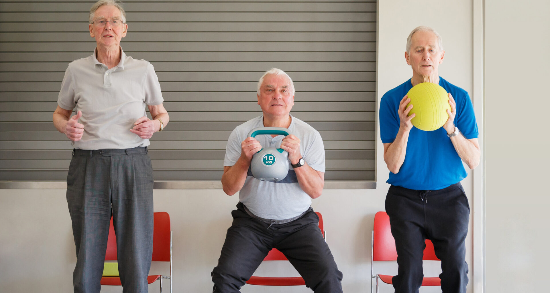 Three men performing chair squats, one is holding a kettlebell, another a yellow ball as they sit back into the chairs