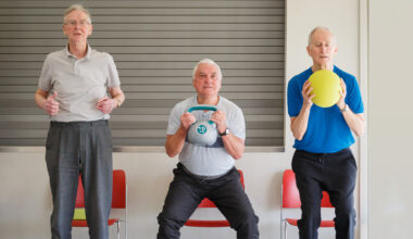 Three men performing chair squats, one is holding a kettlebell, another a yellow ball as they sit back into the chairs