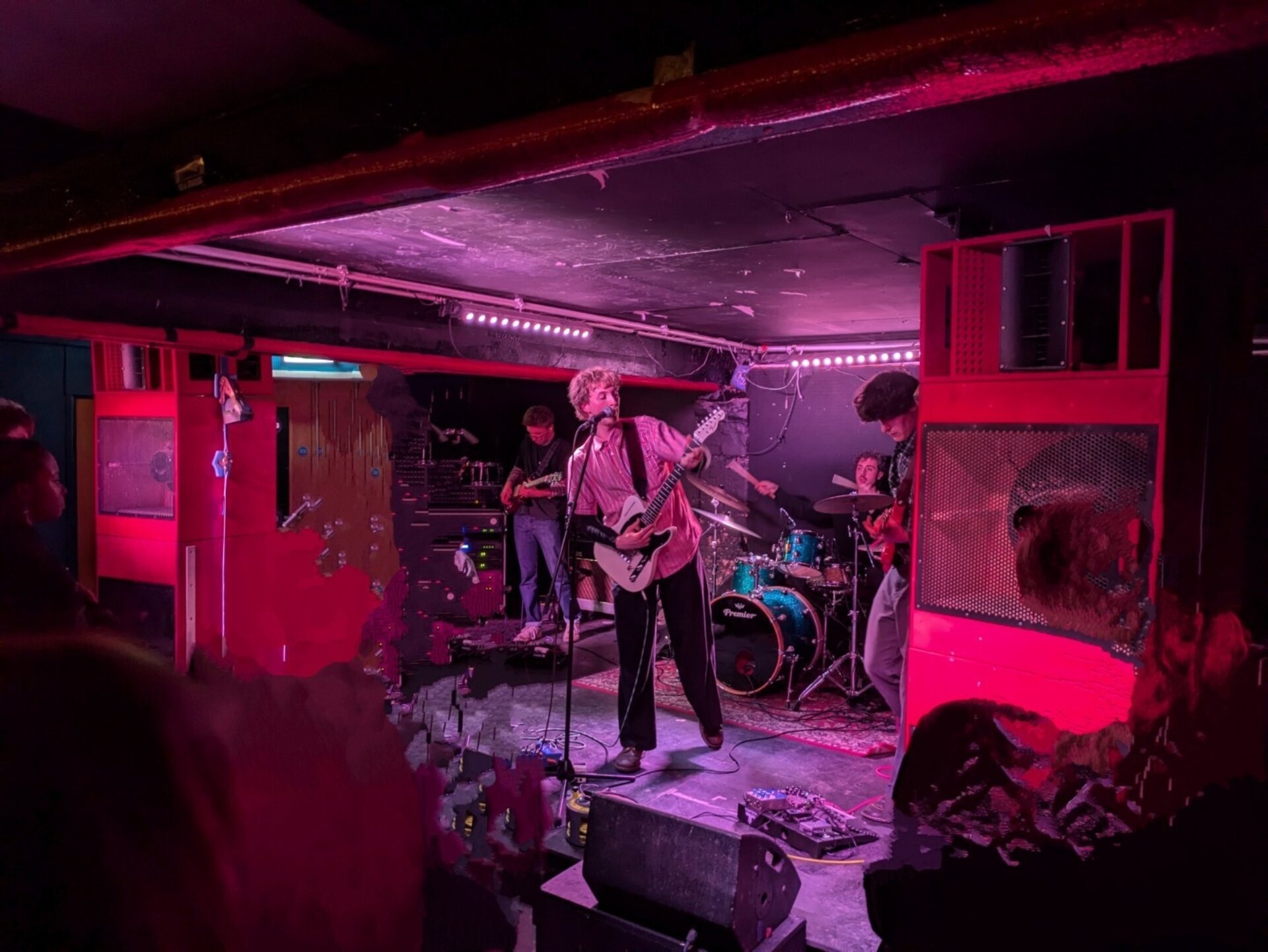A photo of a band, consisting of two guitarists, a drummer and a bass player, performing onstage in a basement setting, with two huge speaker stacks on either side of them and lit by red light