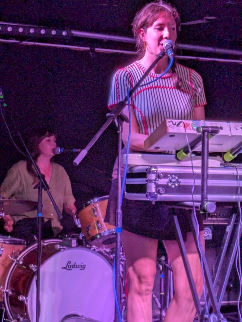 A close-up photo of two musicians performing onstage, one standing up at a synthesiser and the other sat down behind at a drumkit