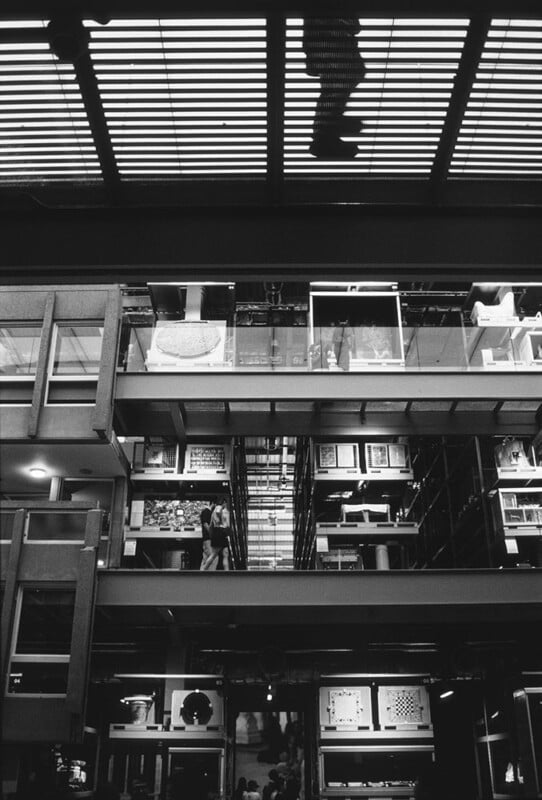 Black and white photo of a multi-story building interior with open walkways and glass walls; a person is visible walking above on a grated floor, casting a shadow below.