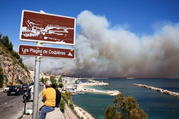 People look on near the Plage des Corbieres, on the outskirts of Marseille, southern France on July 8, 2025, as a smoke from a wildfire rages in the background. The fire that started late on July 8, 2025 morning in Les Pennes-Mirabeau (Bouches-du-Rhone), a town bordering Marseille, has entered France's second-largest city, where some residents were urged to shelter in place, according to a warning message at 4:00 PM from the prefect of Bouches-du-Rhone. (Photo by Clement MAHOUDEAU / AFP) (Photo by CLEMENT MAHOUDEAU/AFP via Getty Images)