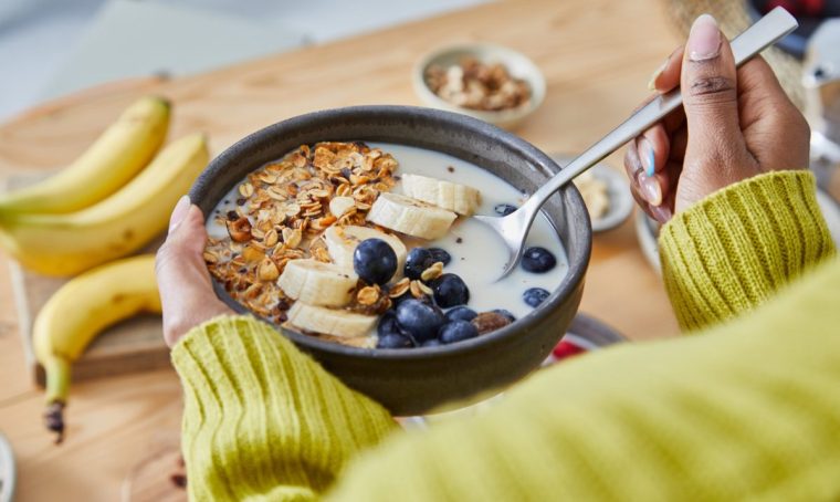 Young adult girl making oatmeal for breakfast, with fresh organic fruit, nuts and grains, at a wooden table in a modern white kitchen, representing a healthy lifestyle, an image with a copy space