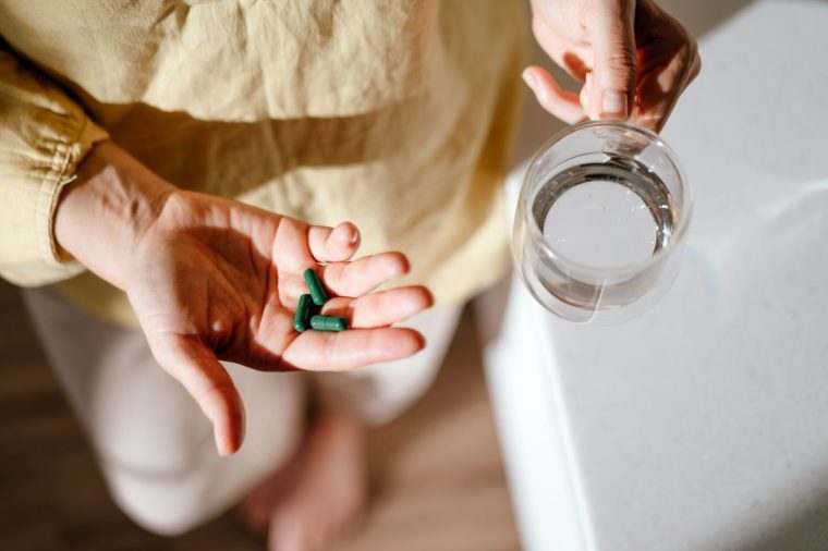 A woman standing at a table with green pills in her hand. Taking vitamins, macro elements, chlorophyll, spirulina and bioactive supplements to maintain health. Prevention of colds during an epidemic. The first symptoms of incipient menopause are hypotension or hypertension, hot flashes.