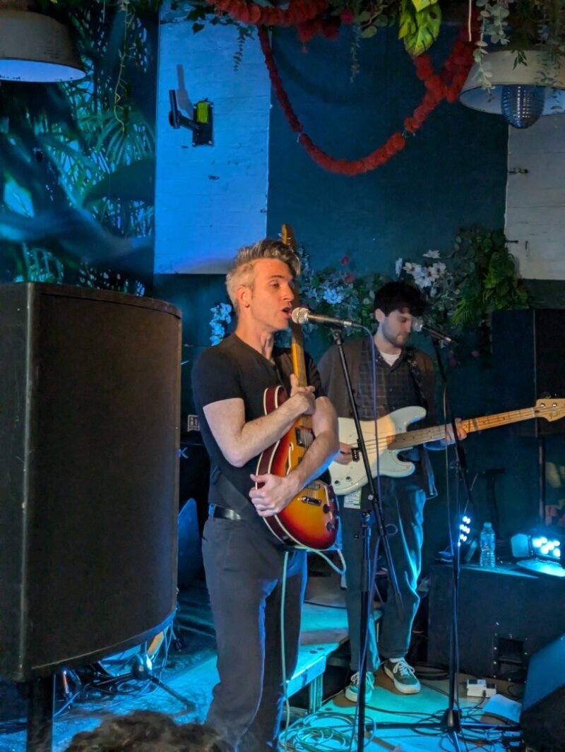 A close-up photo of a guitarist and bassist performing onstage, with the guitarist singing into a microphone
