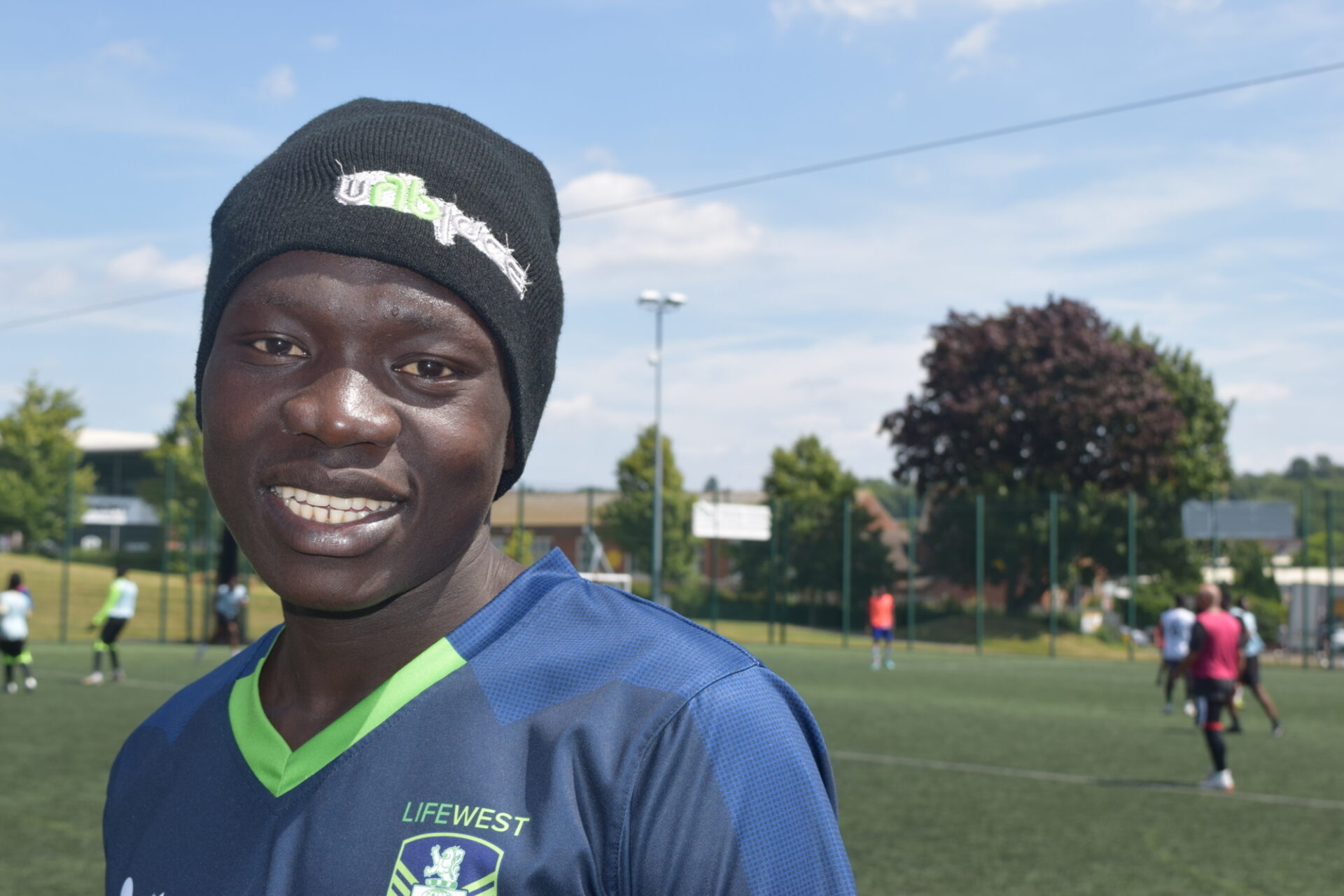 A young man in a football shirt and a beanie hat smiles at the camera while people play football on the pitch behind him.