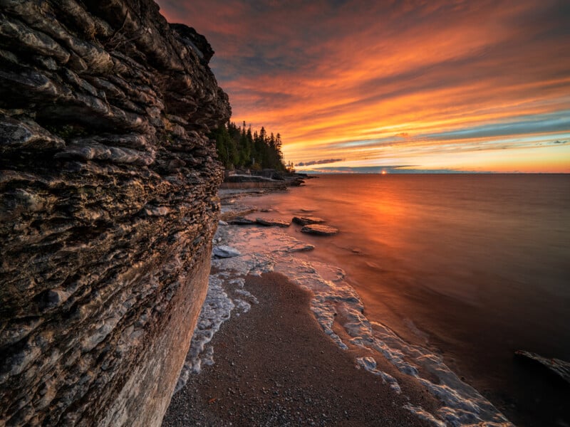 A dramatic sunset over a rocky lakeshore, with layered cliffs on the left, smooth water reflecting orange and red hues, and a line of trees silhouetted against the colorful sky.