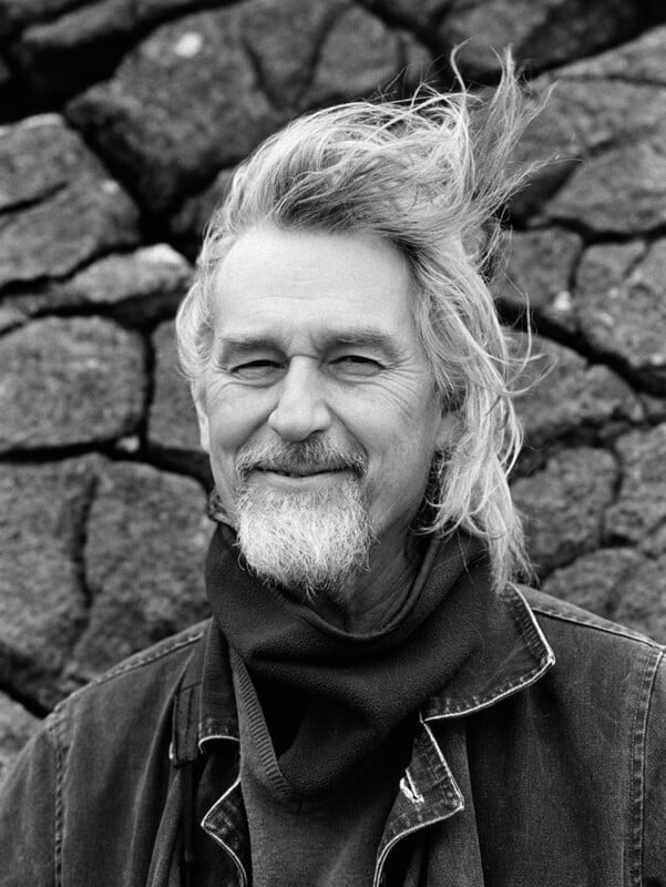 A smiling older man with windswept gray hair and a beard stands in front of a stone wall, wearing a scarf and denim jacket. The photo is in black and white.