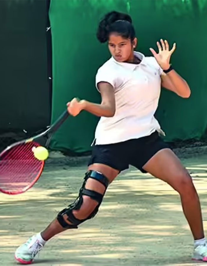 Young female tennis pro wearing knee brace hitting a backhand shot on an outdoor clay court during practice.