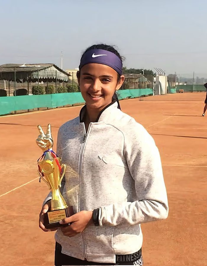 Young female tennis pro smiling on clay court holding a trophy, highlighting male ego controversy in sports.