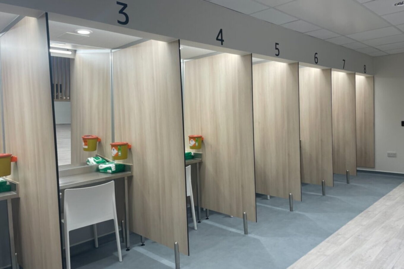 A photo of a room with a series of booths, numbered 3 to 8, each with a mirror, a small table, a chair, and some equipment for safely injecting drugs