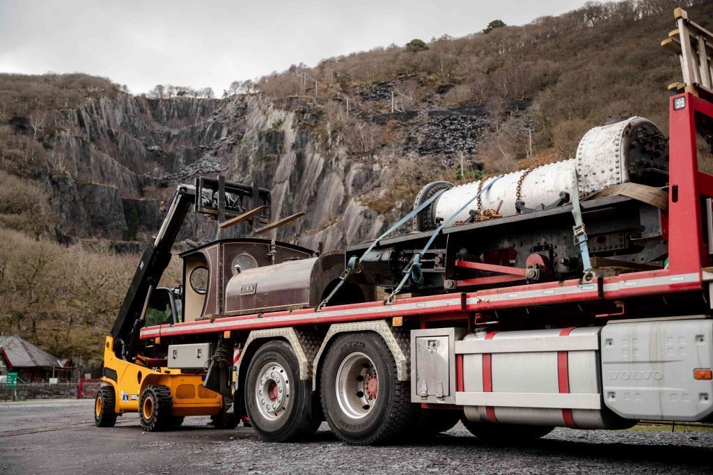A forklift loads a disassembled steam locomotive onto a flatbed truck, set against a mountainous, rocky landscape with sparse trees and overcast skies.