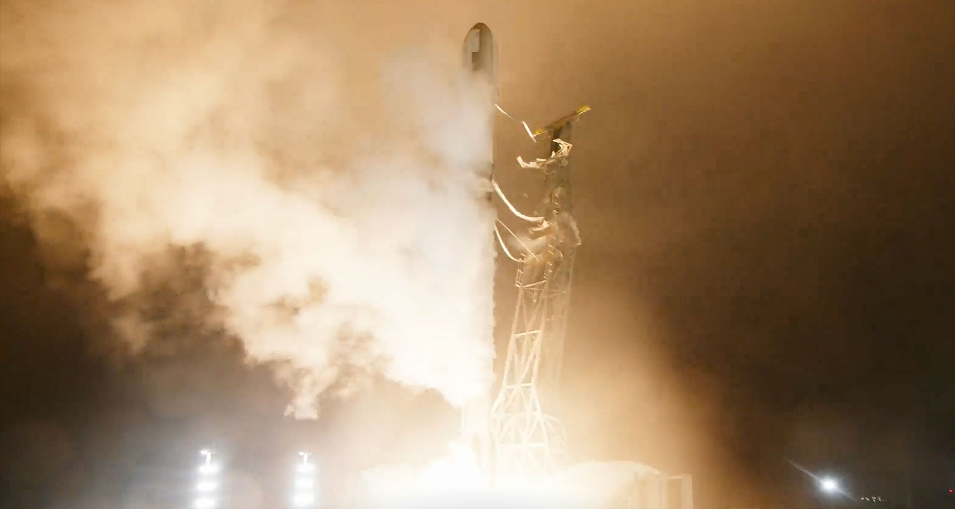 a white and black rocket lifts off at night, its engine thrust lighting up its launch pad and surrounding area