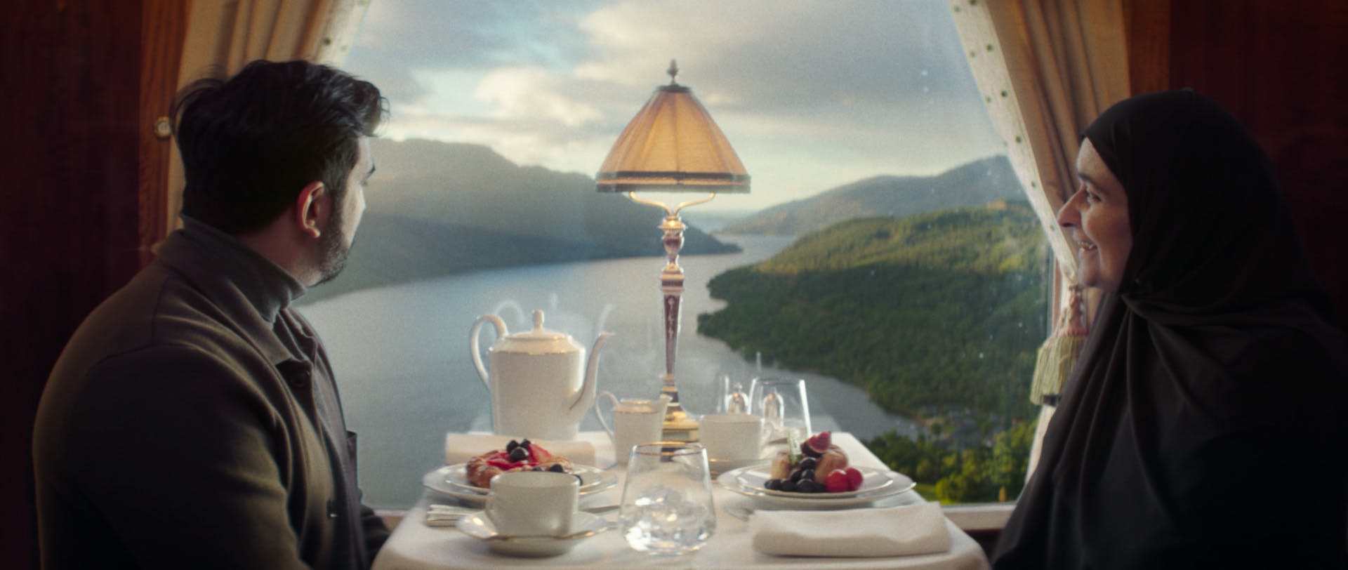 Still from a VisitBritain ad showing two people sat at a table with afternoon tea looking out of a train window onto Loch Lomond