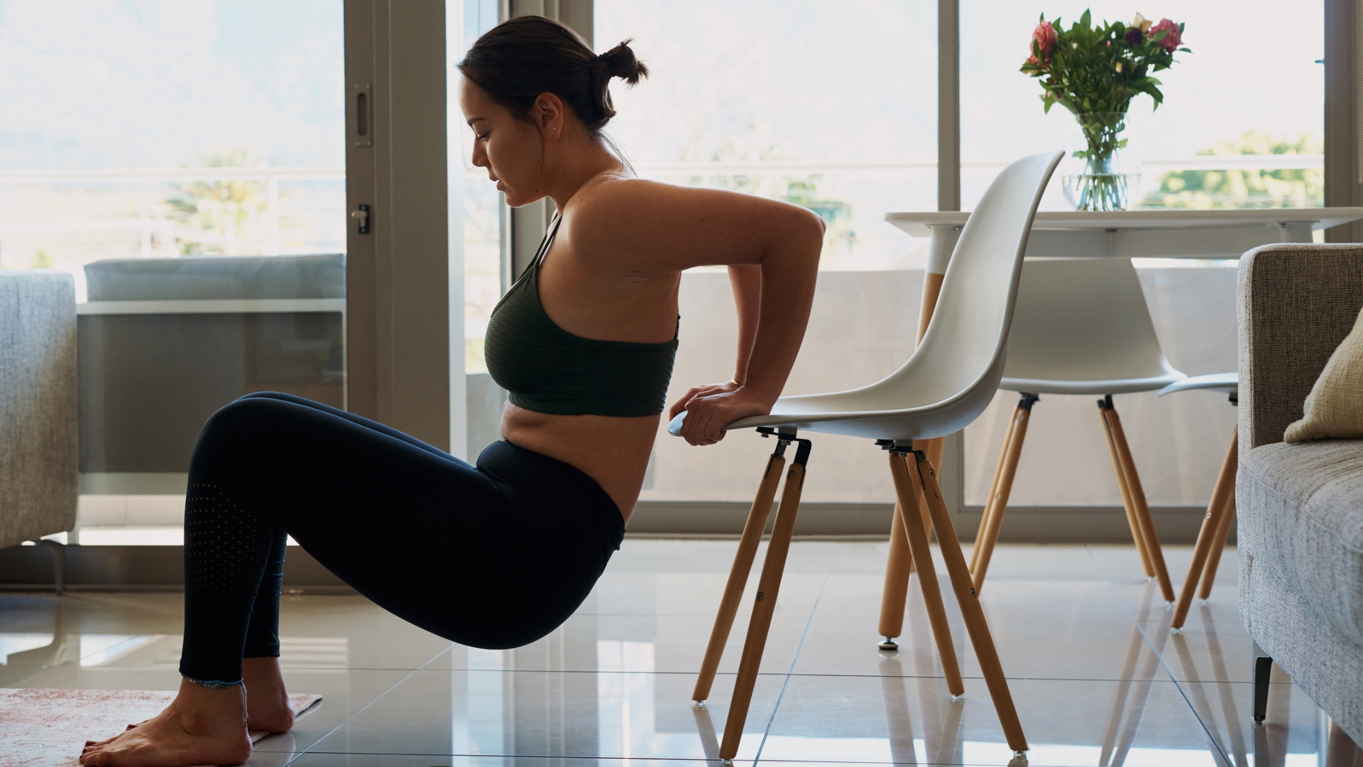 woman wearing dark crop top and leggings performing a triceps dip in a living room setting on a white chair, facing sideways to the camera.