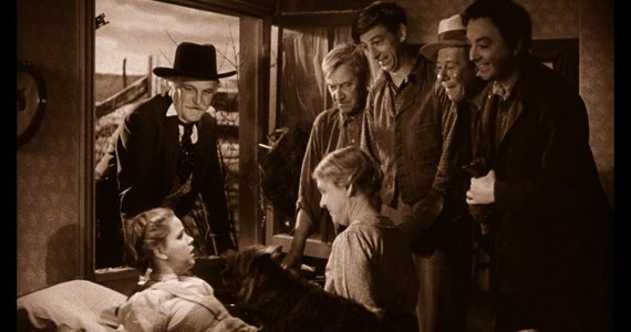 Scene from a film depicting a group of men surrounding a young girl lying in bed.  A man in a hat stands behind her.