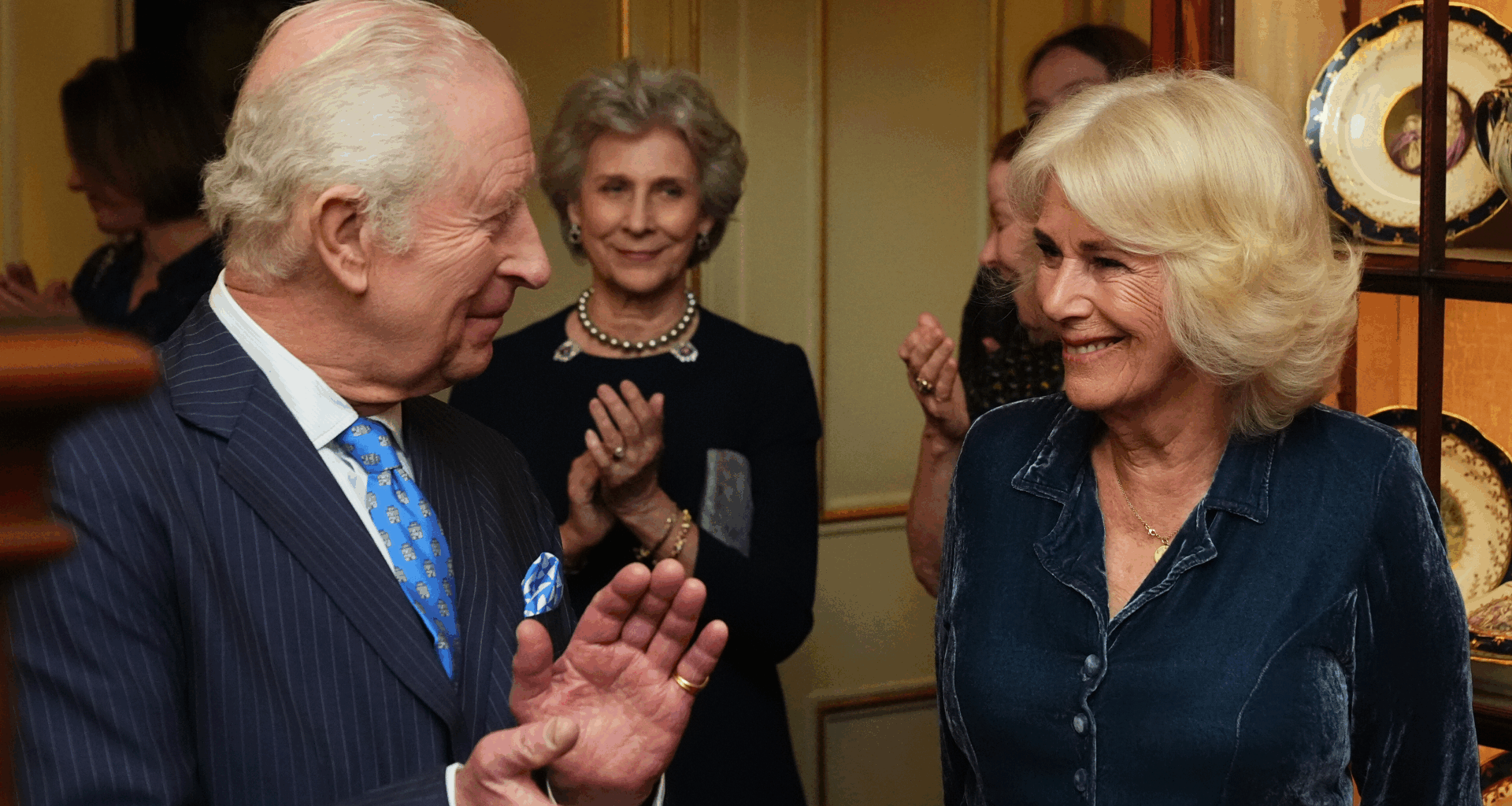 King Charles clapping and smiling at Queen Camilla, who is dressed in a blue dress