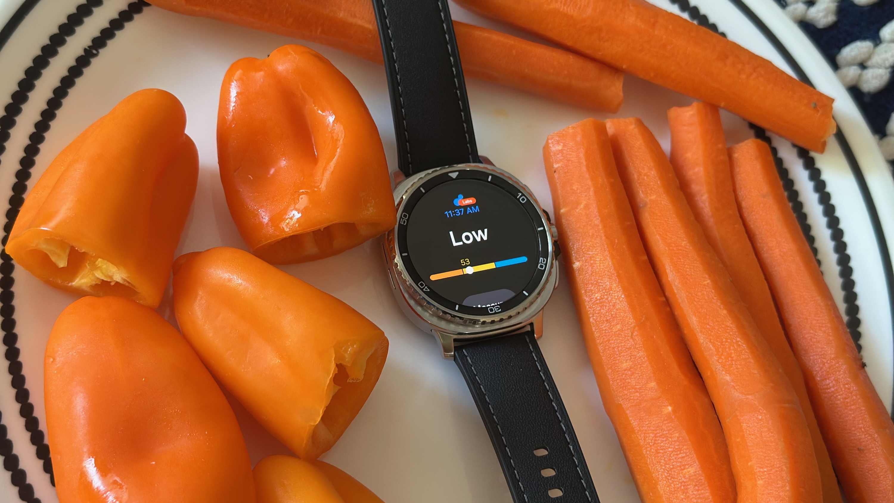 The Samsung Galaxy Watch 8 Classic sitting on a plate surrounded by bell peppers and carrots, the display showing the Antioxidant widget with a 