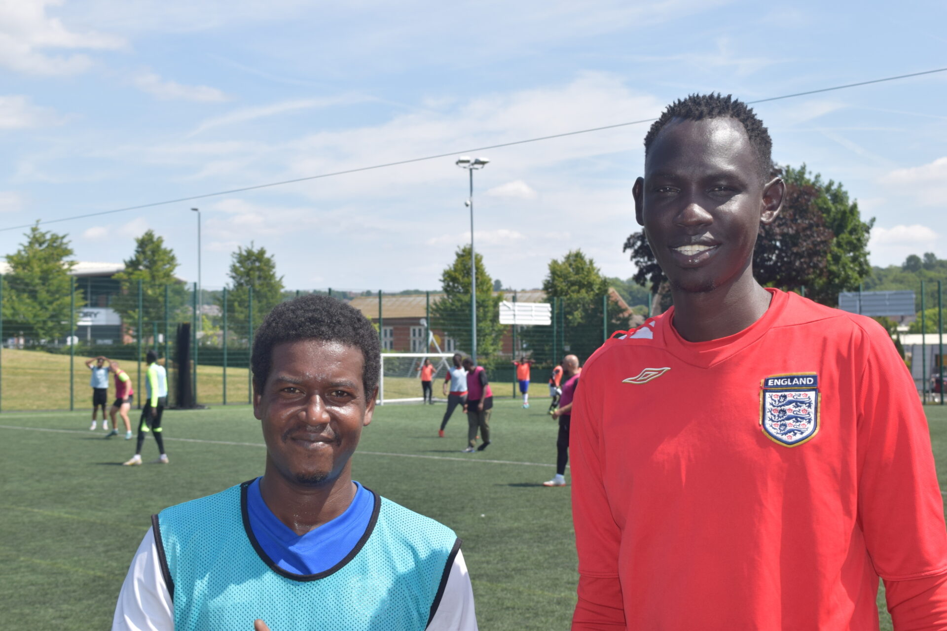 Two middle aged men, one tall and one shorter, are standing on a football pitch looking at the camera, with a game happening behind them.
