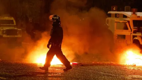 Reuters A police officer in full protective gear, including a helmet. They are walking past two areas of fire and a police land rover can be seen in the background. 