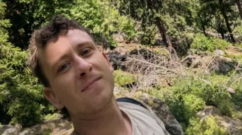 Submitted A man with short brown hair which is slightly curly on top taking a selfie surrounded by rocks, trees and general greenery. He is wearing a beige top and a black backpack.