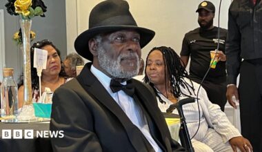 Guy Reid-Bailey looks off towards the right of the image, wearing a black suit with white shirt and black hat. In the background other party goers are visible