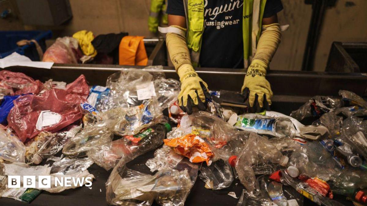 A conveyor belt with dozens of crushed and squashed soft plastics, including bottles and plastic bags. There is a person standing above it with their hands on some of the plastic, wearing thick yellow gloves and a hi-vis jacket