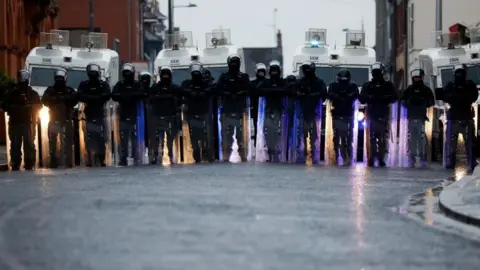 Reuters A line of riot police in front of police land rovers. They are wearing full protective gear, including helmets and protective shields. 