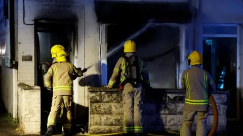 Reuters Four firefighters in high vis uniforms and yellow hard hats have their backs to the camera. They are hosing down a white house. The house is charred black above the front door and living room window. There is smoke coming from the window which has been smashed.