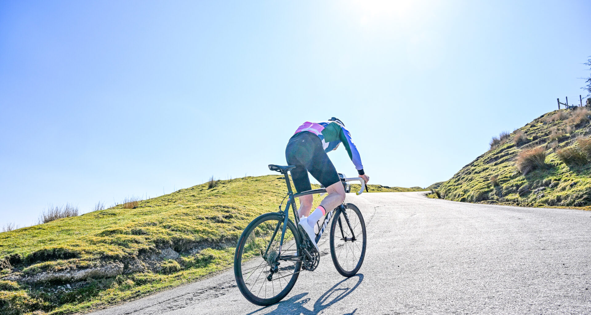 A rider climbing a hill, blue kit, blue sky, glaring sunlight, with the sun directly overhead and almost in shot