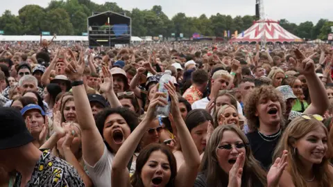 BBC/Oli Constable A crowd stands in front of a stage, some are clapping, others have their hands in the air and are singing along.