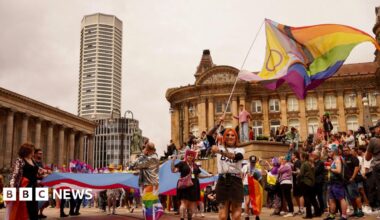 A woman with orange hair waves a rainbow flag in a wide shot Chamberlain Square Birmingham. She is wearing a white t-shirt and black mini skirt. Behind her are crowds of people wearing rainbow accessories. Birmingham's council house can be seen to the left and the Octagon tower behind.