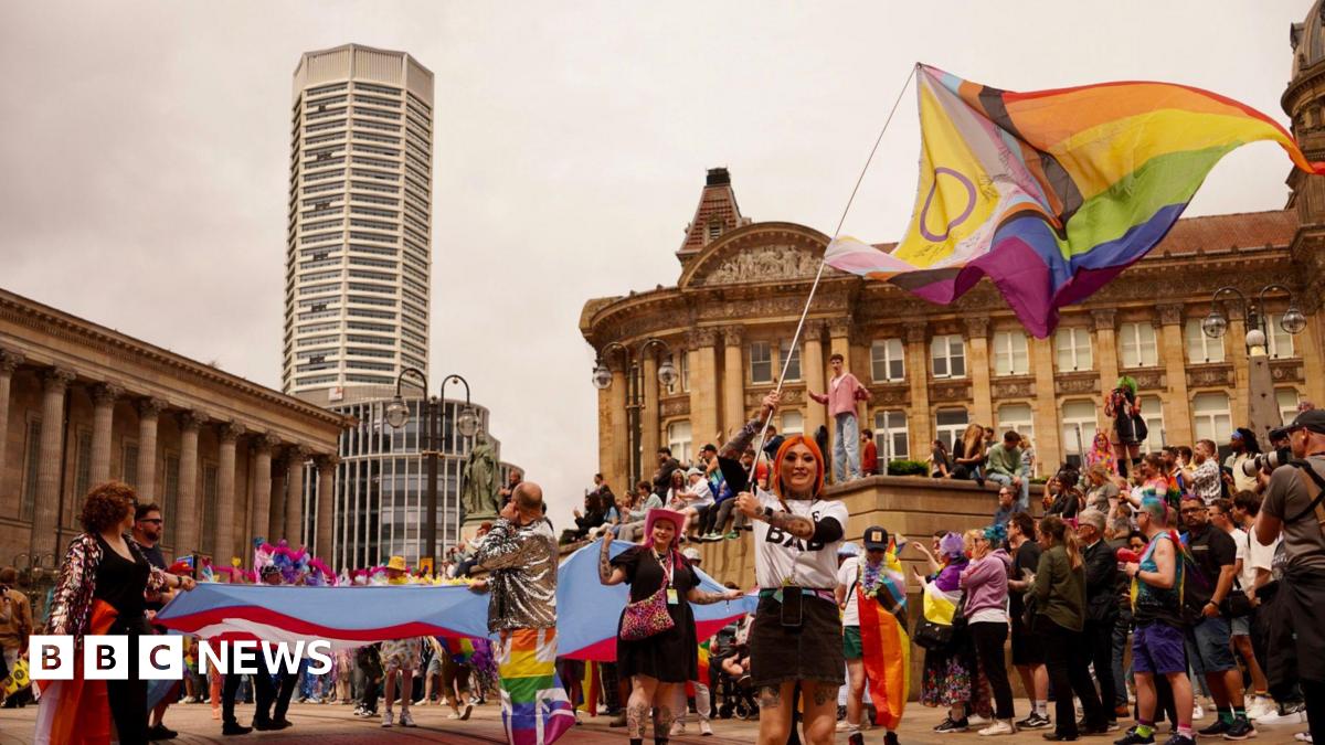 A woman with orange hair waves a rainbow flag in a wide shot Chamberlain Square Birmingham. She is wearing a white t-shirt and black mini skirt. Behind her are crowds of people wearing rainbow accessories. Birmingham's council house can be seen to the left and the Octagon tower behind.