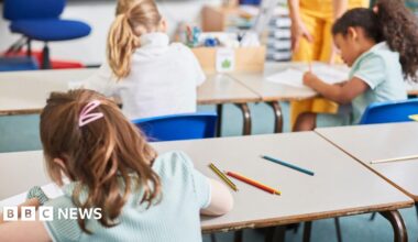 Three school girls have their backs to the camera. They are using pencils to write on pieces of white paper. The child closest to the camera has brown hair and two pink clips in her hair. On the desk beyond her are two children. The one on the left has blonde hair in a ponytail. The girl to the right has black hair in a ponytail. They sit on blue chairs at white desks.
