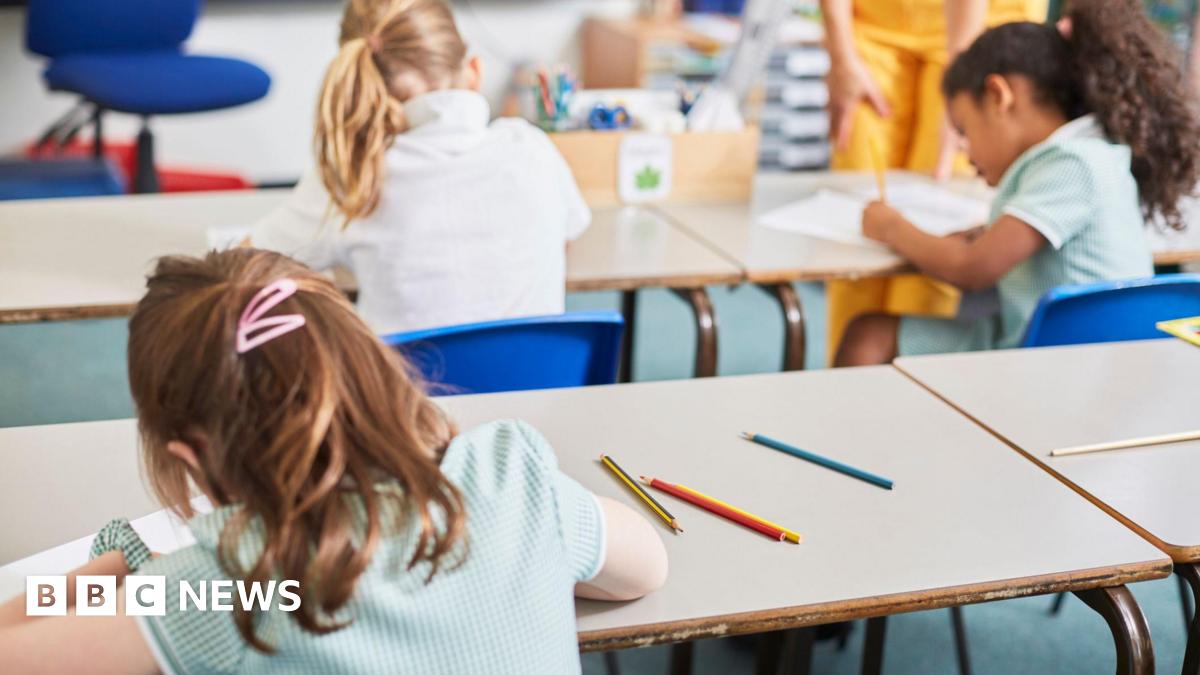 Three school girls have their backs to the camera. They are using pencils to write on pieces of white paper. The child closest to the camera has brown hair and two pink clips in her hair. On the desk beyond her are two children. The one on the left has blonde hair in a ponytail. The girl to the right has black hair in a ponytail. They sit on blue chairs at white desks.