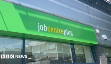 The Jobcentre Plus sign, with white and yellow lettering on a green background, with a window underneath reflecting the street opposite