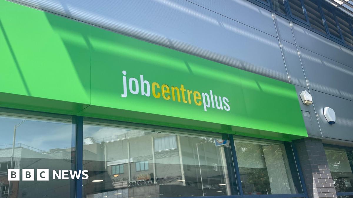 The Jobcentre Plus sign, with white and yellow lettering on a green background, with a window underneath reflecting the street opposite
