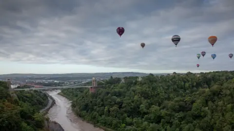 Getty Images Scenic view of Clifton Suspension Bridge crossing the River Avon gorge surrounded by green forests. Several hot air balloons float in the sky above. In the background, a cityscape and distant hills are visible under a cloudy sky