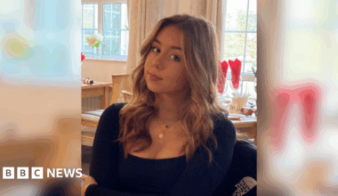 Jessica with brown highlighted hair in loose, wavy curls. She is wearing a black square-necked top.  She is sitting down in a restaurant with brown wooden tables behind her.