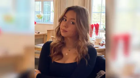 Glasser family Jessica with brown highlighted hair in loose, wavy curls. She is wearing a black square-necked top.  She is sitting down in a restaurant with brown wooden tables behind her.