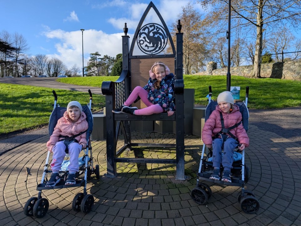 Three young girls, one sitting on an ornate chair and two in strollers, at a park.