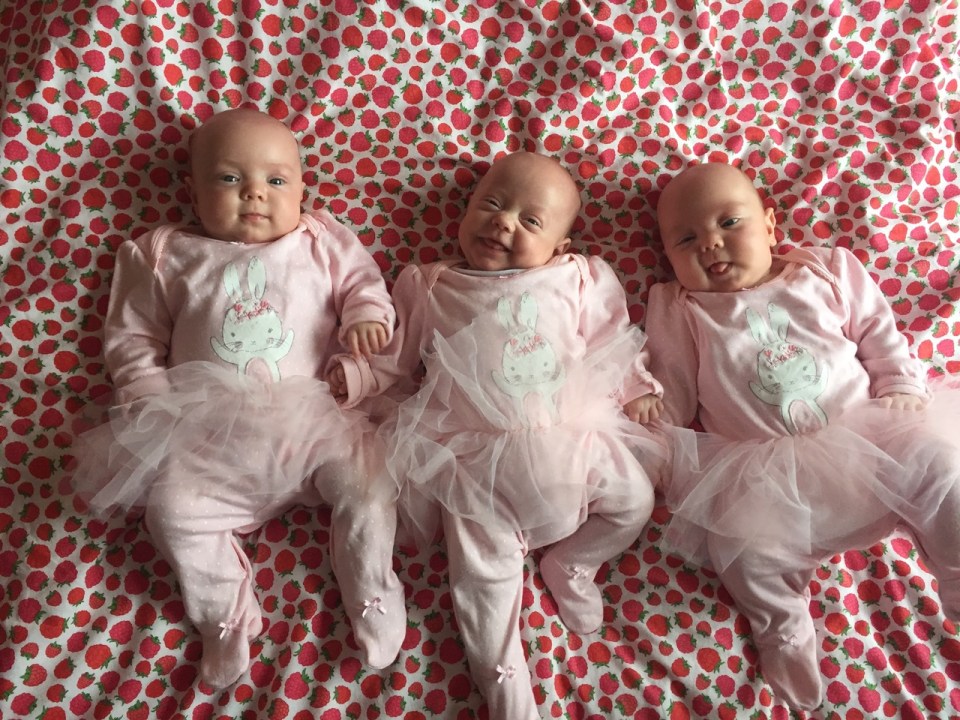 Three baby girls in pink outfits lying on a strawberry-patterned blanket.
