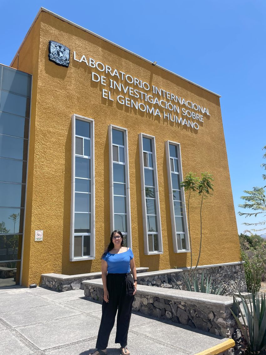Alejandra Medina-Rivera in front of a yellow building bearing the sign ‘LABORATORIO INTERNACIONAL DE INVESTIGACIÓN SOBRE EL GENOMA HUMANO’ and the emblem of the National Autonomous University of Mexico.