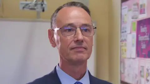 BBC Mark Browne  has receding grey/brown hair and brown eyes and glasses standing in front of a a school white board and pink billboard. He is wearing a navy suit, white tie and blue shirt and has a neutral expression.